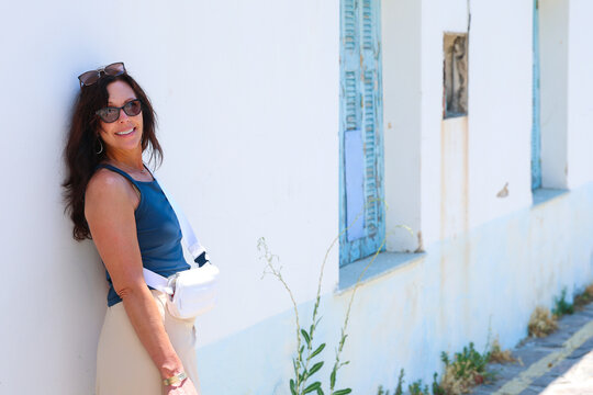 A smiling woman wearing sunglasses leans against a white wall with blue shutters in Plaka, Milos, Greece, enjoying the warm Mediterranean sunlight and relaxed island atmosphere.