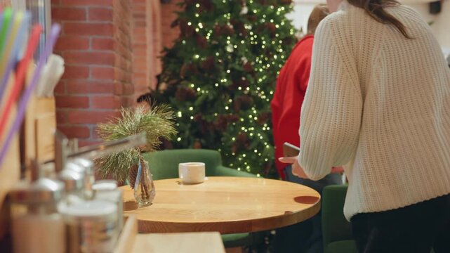 Cozy cafe table near Christmas tree, patron in knit sweater pulls chair, sets ceramic mug, surveys warm holiday decor, brick wall, greenery centerpiece, soft fairy lights, peaceful morning atmosphere