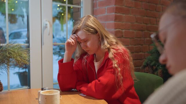 White woman in bustling cafe by window, wearing red jacket, rubbing forehead and checking phone, tense conversation with friend across table, coffee cups on wooden table, winter light through glass,