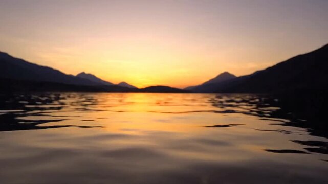Mountains rise above a tranquil lake as the golden sun sets on the horizon during a peaceful evening.