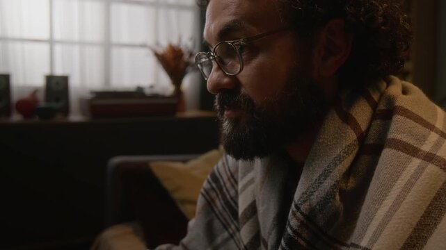 Middle aged Caucasian man wrapped in blanket drinking hot tea and using remote control while watching television. Perfect for home entertainment and streaming service advertisements