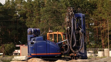 Niebieska wiertnica gąsienicowa z hydraulicznym osprzętem stoi na placu budowy. W tle widać las, kontenery oraz elementy betonowe. Maszyna jest przygotowana do specjalistycznych prac ziemnych. © tomo81