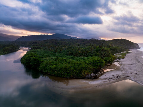 Aerial view of Parque Nacional Natural Tayrona where a river meets the sea, surrounded by lush tropical forest and sandy beaches under a dramatic sunset sky in Magdalena, Colombia.