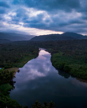 Aerial view of Parque Nacional Natural Tayrona featuring a winding river flowing through dense tropical forest with mountains under a dramatic cloudy sky in Magdalena, Colombia.
