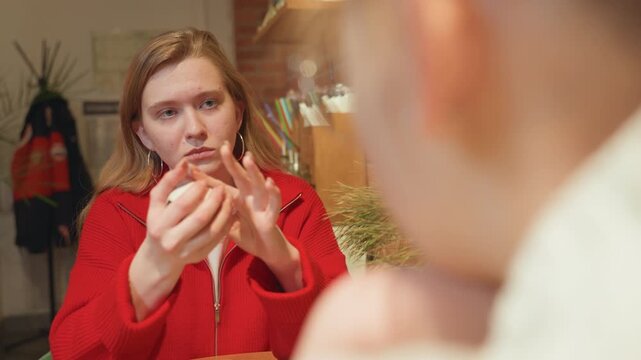 Woman in red jacket holding cup at cafe table, serious listening expression with hands clasping mug, indoor coat rack and warm lighting in background tense but quiet exchange, young professional mood