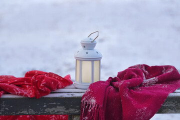 A white lantern with stars and a red plaid blanket sitting on a snowy wooden bench outdoors. Winter, Christmas © Natalia