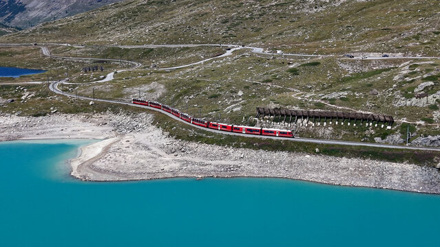 Aerial view of the red Bernina Express train traveling along the tracks beside the turquoise waters of Lago Bianco in the Swiss Alps near Poschiavo, Grisons, Switzerland.