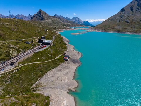 Aerial view of the Bernina Express train traveling along the turquoise waters of Lago Bianco in the Bernina Pass, surrounded by rugged alpine mountains in Poschiavo, Grisons, Switzerland.