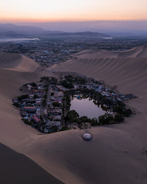 Aerial view of Huacachina Oasis surrounded by towering sand dunes and palm trees at sunset in Huacachina, Ica, Peru.