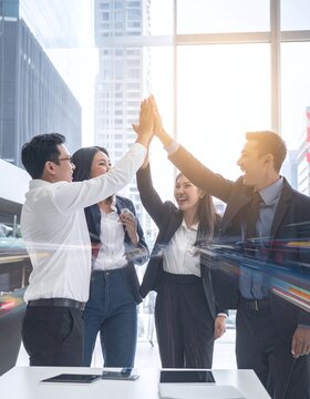 A cheerful Asian business team celebrating corporate success with a highfive in a modern skyscraper office, inspiring unity, teamwork, and shared professional achievement.