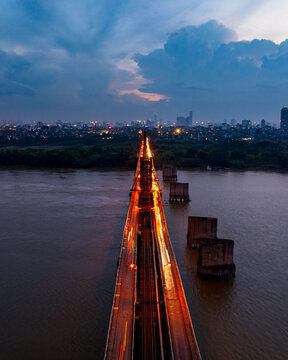 Aerial view of Long Bien Bridge illuminated at night with light trails from traffic crossing the Red River, set against the city skyline of Hong Ha, Ha Noi, Vietnam.