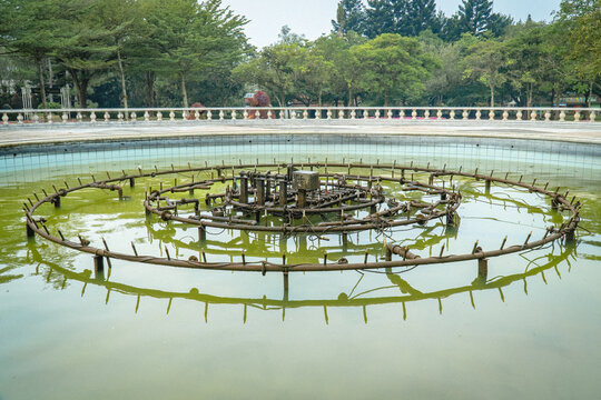 Circular fountain mechanism installed in a pond, surrounded by calm water and trees in a landscaped public park.