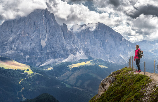 Woman hiker in red jacket with backpack standing on cliff edge viewpoint looking at mountain peak in Dolomites, Italy. Alpine summer landscape with green valley, trail, fence and dramatic cloudy sky