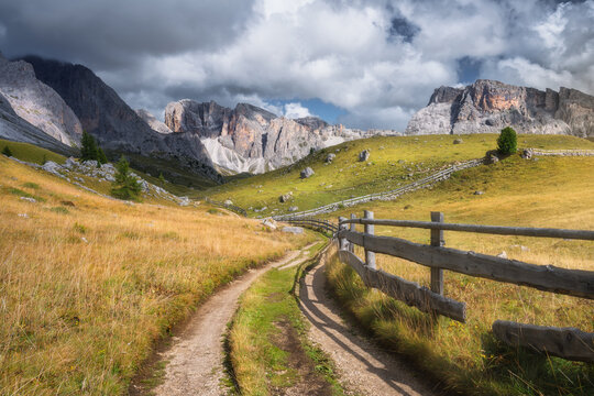 Dirt road with wooden fence leading to mountain peaks in Dolomites, Italy. Autumn alpine landscape with gravel path, green grass meadow, dramatic dark clouds and sunlight on rocky cliffs at sunset