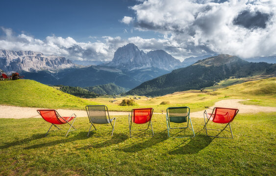 Empty deck chairs on green grass meadow facing mountain range view in Dolomites, Italy. Summer alpine resort landscape with colorful lounge armchairs on lawn against cloudy sky and rocky peaks. Alps