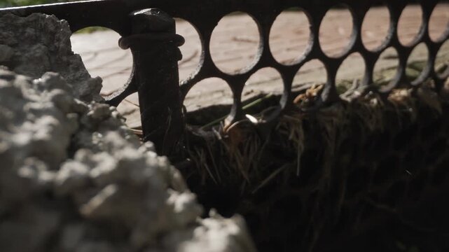 Macro cement mound with gritty powder and coarse aggregate in foreground against circular iron fence bokeh. Lowangle tactile closeup shows crumbly particles, porous surfaces, shadowed cavities,