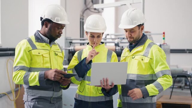 Group of factory engineers wearing safety jackets and helmets discussing project. Female holding laptop showing data while colleague using tablet. Team collaborating in production facility.