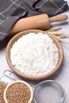 Wheat flour in bowl, grains, spikes, rolling pin and sieve on grey table, above view