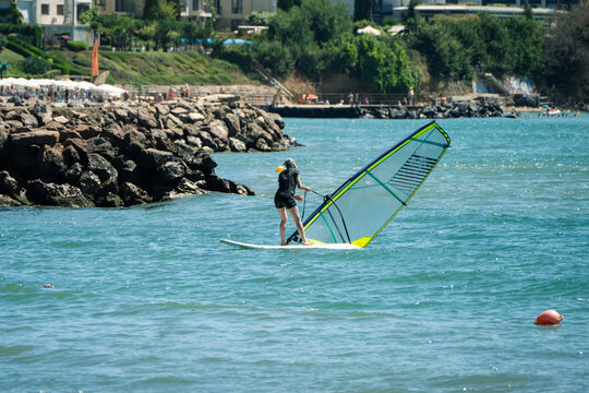 A windsurfer balances on their board while pulling up a yellow and transparent sail in bright blue water near a rocky, tree-lined coastal resort.