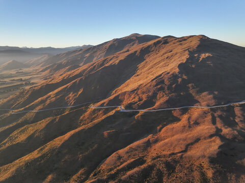 Aerial view of Crown Range Road Scenic Lookout winding through the rugged mountains of the Otago Region during golden hour, Roaring Meg, Otago Region, New Zealand.