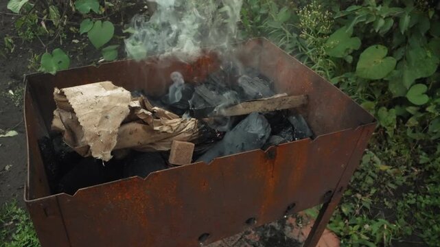 Macro closeup charcoal smoking with thick plume rising, intense ember glow and swirling vapor, oxidized steel grill edges, active combustion texture, leafy wild background, cinematic dramatic mood