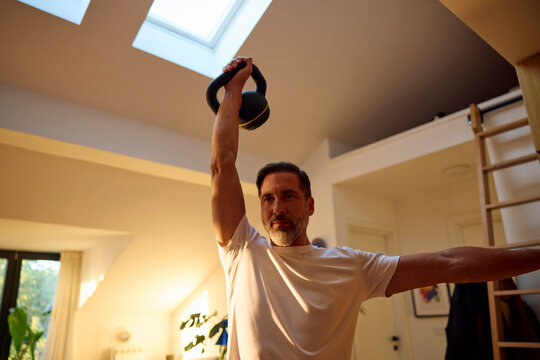 A man exercises in the living room with a kettlebell in hand