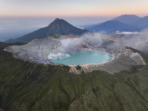 Aerial view of Ijen crater lake with its turquoise water and volcanic steam rising from the rugged rim, surrounded by lush green slopes in East Java, Indonesia.