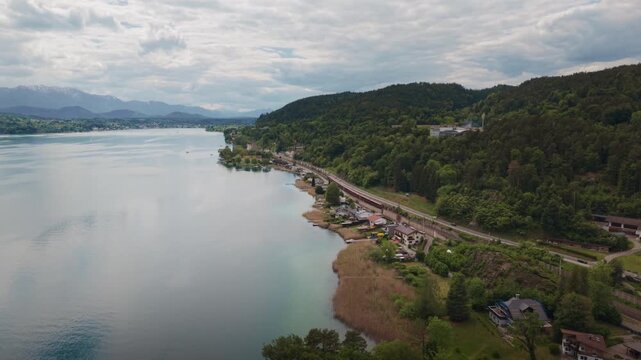 Lakeside Railway Journey: An aerial view captures a train track winding along the serene shore of a vast lake, bordered by lush green forest and distant rolling hills under a cloudy sky.