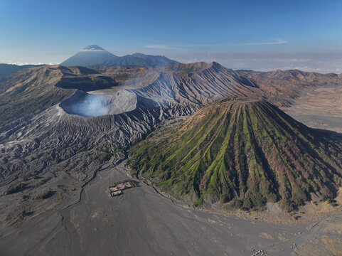 Aerial view of Mount Bromo, showing the volcanic crater, lush green slopes of Mount Batok, and the vast sandy caldera under a clear sky in Tosari, East Java, Indonesia.