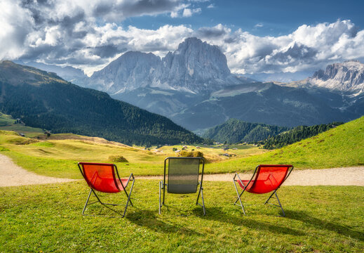 Empty deck chairs on green grass meadow facing mountain range view in Dolomites, Italy. Summer alpine resort landscape with colorful lounge armchairs on lawn against cloudy sky and rocky peaks. Alps
