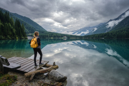 Woman hiker with yellow backpack standing on wooden pier at alpine lake in Dolomites, Italy. Mountain landscape reflected in calm water with forest and cloudy sky on overcast summer rainy day. Travel