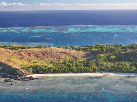 Aerial view of Mana Island featuring a pristine white sand beach, lush green hills, and a vibrant turquoise lagoon with coral reefs and sailboats in Western Division, Fiji.