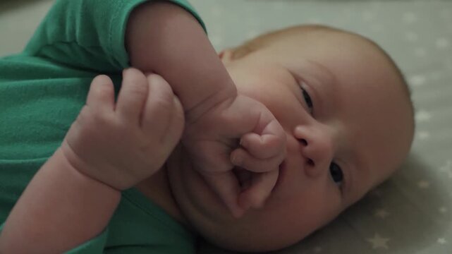 Close up of newborn lying on soft patterned blanket, both hands raised near mouth while fingers touch lips and gentle sucking motion shows self-soothing, calm comfort, and tender warmth