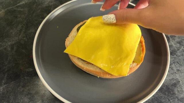 Woman placing a square slice of yellow cheese onto a burger bun, homemade burger preparation, cooking process, fast food.