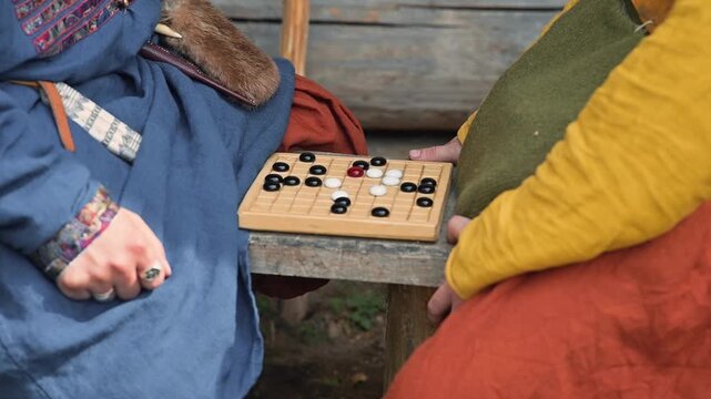 Two players, one wearing a blue garment and the other in a yellow and green outfit, focus on a wooden game board set on a table