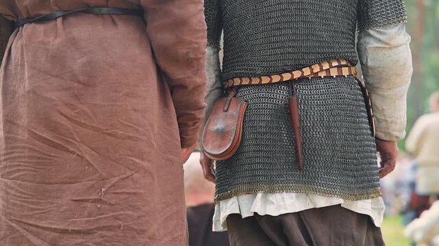 Two male reenactors dressed in medieval tunics and chainmail stand in a grassy area. They carry tools and pouches at their sides, highlighting crafts and daily life from the early Middle Ages