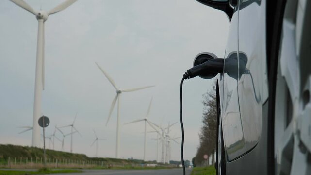 Electric car plugged into roadside charger near row of wind turbines beneath overcast sky, with cable and connector engaged to vehicle port, highlighting clean transport, power and sustainability