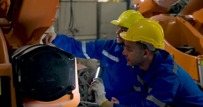 Two adult male technicians crouch beside industrial robotic arm, using flashlight and tool to inspect mechanical joint while diagnosing equipment condition during factory maintenance and repair.