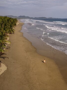 Aerial view of San Miguel beach with a person walking along the sandy shore, lush palm trees, rolling waves, and distant mountains in San Miguel, Provincia de Guanacaste, Costa Rica.