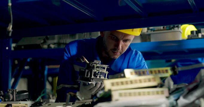 Adult male technician inspects electrical control component inside industrial machinery, lifting wired module for close examination during diagnostics and preventive maintenance in factory workshop.