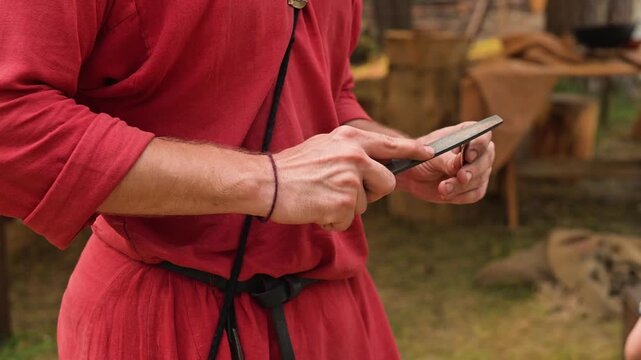 A person in a red tunic carefully shapes a wooden tool . The setting features a rustic backdrop with tents, suggesting an early medieval camp atmosphere