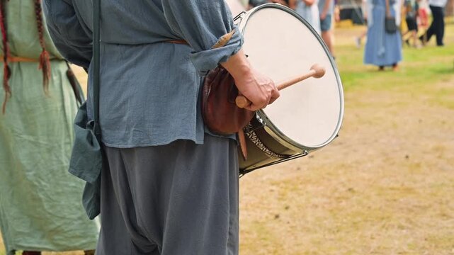A male drummer stands in a field, holding a drum with a stick. He is dressed in loose blue clothing and is part of a larger group of people wearing historical attire engaged in activities