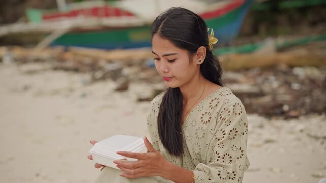 Young asian woman beach eating takeout box, sitting by shoreline, unfolding container, tasting snack with quiet smile, traveler and storyteller roles, braid with flower, boat in background, sandy