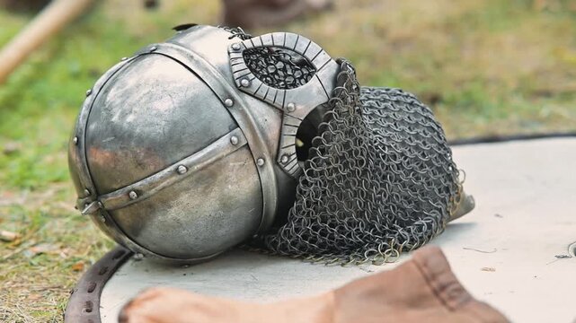 A chainmail coif and an armored helmet lie on the ground . The setting features a grassy field that indicates a historical reenactment atmosphere