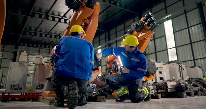 Two adult male technicians crouch beside industrial robotic arm, checking lower mechanical sections with inspection light and discussing equipment condition during collaborative factory maintenance.
