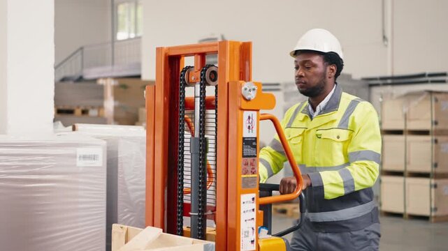 African American male factory worker wearing reflective jacket and white helmet pushing orange lifter with determination. Focused expression while moving materials in spacious warehouse.