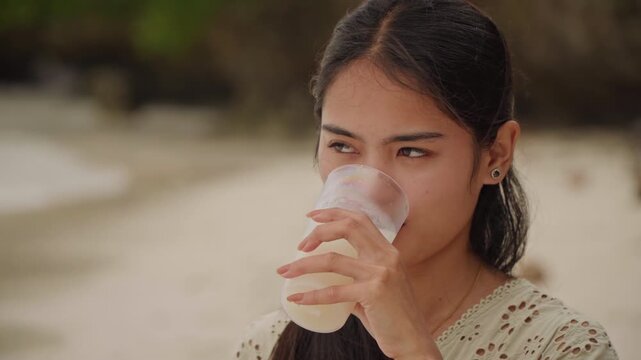 Sipping coconut water at beachside moment. Closeup of woman enjoying fresh coconut beverage at shore. Expressive face captures fleeting joy while drinking coconut water on tropical beach