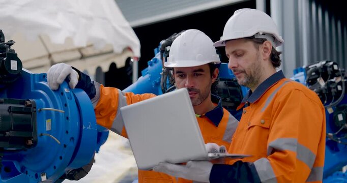 Two adult male technicians examine industrial robotic arm joint, pointing at machinery and reviewing laptop data during collaborative diagnostics and maintenance inside automated factory workshop.