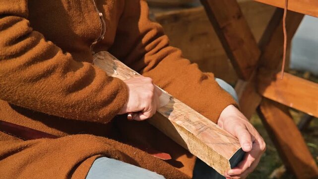 A person sits on a wooden bench, using a tool to shape a piece of wood. The individual appears to be in their twenties or thirties, dressed in simple clothing typical of the early Middle Ages