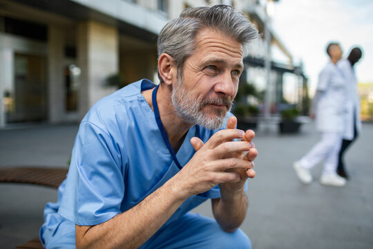 Thoughtful male doctor in scrubs sitting outside hospital